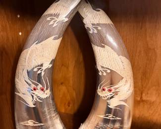 Hand-Carved Dragon Horn Sculptures - Pair. Intricate hand-carved bovine horn sculptures featuring detailed dragon motifs. This pair has polished natural horn surfaces with cream-colored carvings and red bead eyes. Each horn is mounted on a dark-stained wooden base.