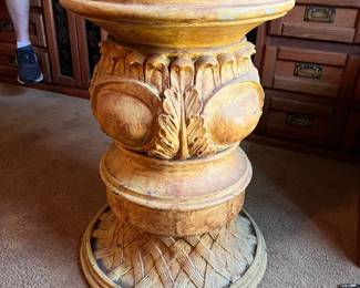 Ornate pedestal table is made of a cast resin or composite material with a distressed yellow and tan finish. It has a circular top and a base featuring intricate acanthus leaf carvings and a woven lattice pattern at the bottom.