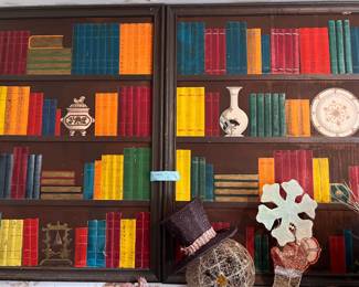 Vintage Trompe L'oeil Bookshelf Wall Art Panels - Set of 2. Hand-painted wooden wall panels with a library bookshelf motif. These rectangular frames feature a dark brown finish and depict rows of colorful books, vases, and decorative plates in a trompe l'oeil style. The set includes two matching panels with integrated wooden frames.