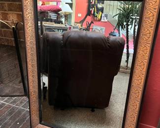 Decorative Copper-Toned Framed Wall Mirror. Ornate wall mirror with a copper-toned resin or wood frame. The frame has a repeating scrollwork pattern and a dark inner border. The glass is clear and rectangular.