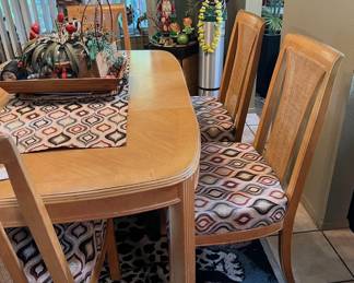 Sturdy wood dining set includes an oval-shaped light wood table, two leaf extensions for larger gatherings, and five matching chairs. The chairs have cane-inlay backs and newly upholstered seats with a geometric pattern in shades of red, grey, and black. The table has a ribbed pedestal base and a smooth wood grain finish.