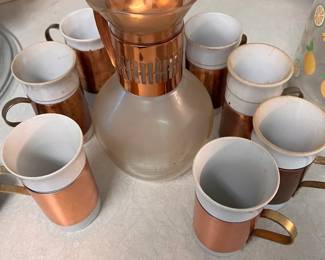 Vintage Copper and Glass Coffee Carafe Set with 8 Mugs. Vintage coffee service set includes one glass carafe with a copper-toned metal collar and lid, and eight matching ceramic mugs with copper-toned metal sleeves and handles. The carafe is clear glass with a copper-colored metal trim. The mugs are white ceramic with copper-colored metal accents.