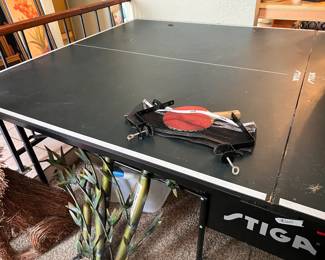 Durable STIGA ping pong table has a dark green playing surface with white boundary lines. The set includes a black STIGA net with metal screw-on clamps and a red and black STIGA paddle with a wooden handle. The table is a folding model with a black metal frame and wheels for mobility.