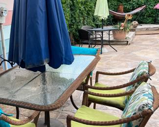 5-Piece Wicker Outdoor Dining Set with Glass Top Table. Durable outdoor dining set includes a rectangular glass-top table with a brown wicker border and four matching wicker armchairs. The chairs have green seat cushions and patterned back pillows in blue, green, and white. The table has a metal frame and a central hole for the umbrella pole. (Umbrella not included)