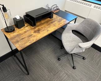 Modern office writing desk featuring a rustic brown wood-grain surface and a black metal frame with built-in lower storage shelves; LumiSource task chair.