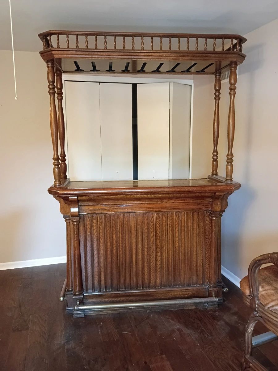 Wooden Home Bar with Overhead Glass Rack and Two Bar Chairs Large traditional-style wooden home bar featuring turned columns, decorative spindle railing, paneled/louvered cabinet doors, a raised serving surface, brass-tone foot rail, and overhead stemware storage slots. Interior cabinets provide additional storage for bottles, glassware, and bar accessories. Includes two matching rattan-style swivel bar chairs with curved arms, padded brown seats and backs, and metal footrest protectors. The set shows visible age and use, including scratches, scuffs, finish loss, upholstery wear, and other blemishes. Contents and decorative items shown are not included. Large and heavy; buyer is responsible for bringing adequate help and equipment for removal. Please inspect all photographs carefully for condition.