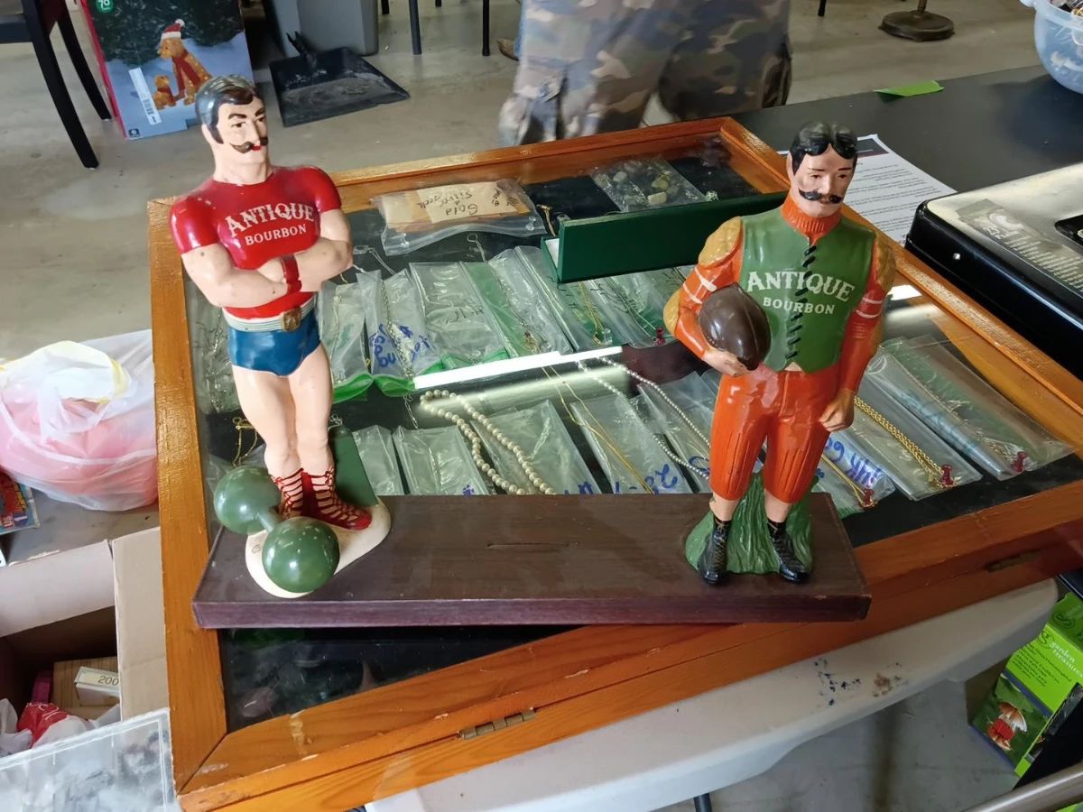 Vintage Antique Bourbon Advertising Figurines Pair of vintage Antique Bourbon advertising display figurines mounted on a wooden base. Features two colorful male figures dressed in old-fashioned athletic attire, each marked “Antique Bourbon.” One figure holds a football, while the other stands beside two balls. Shows visible paint loss, scuffs, and age-related wear. Please review photos carefully for condition and details. Maker, age, and material are unknown.