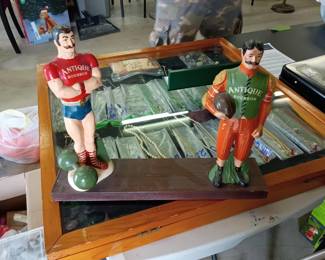 Vintage Antique Bourbon Advertising Figurines  Pair of vintage Antique Bourbon advertising display figurines mounted on a wooden base. Features two colorful male figures dressed in old-fashioned athletic attire, each marked “Antique Bourbon.” One figure holds a football, while the other stands beside two balls. Shows visible paint loss, scuffs, and age-related wear. Please review photos carefully for condition and details. Maker, age, and material are unknown.