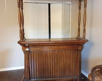 Wooden Home Bar with Overhead Glass Rack and Two Bar Chairs  Large traditional-style wooden home bar featuring turned columns, decorative spindle railing, paneled/louvered cabinet doors, a raised serving surface, brass-tone foot rail, and overhead stemware storage slots. Interior cabinets provide additional storage for bottles, glassware, and bar accessories.  Includes two matching rattan-style swivel bar chairs with curved arms, padded brown seats and backs, and metal footrest protectors. The set shows visible age and use, including scratches, scuffs, finish loss, upholstery wear, and other blemishes. Contents and decorative items shown are not included. Large and heavy; buyer is responsible for bringing adequate help and equipment for removal. Please inspect all photographs carefully for condition.