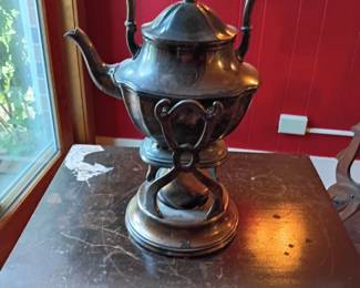 Antique Silver-Plated Tilting Kettle on Warming Stand  Large antique silver-plated hot-water or tea kettle presented on an ornate tilting warming stand. Features a tall swing handle, curved pouring spout, hinged lid with decorative finial, scrollwork supports, round base, and removable burner beneath the kettle. The tilting design allows the kettle to pivot forward for pouring while positioned on the stand.  Shows substantial age-related wear, including heavy tarnish, oxidation, scratches, discoloration, plating loss, residue, surface pitting, and other blemishes. Burner and tilting mechanism have not been tested. Maker and exact age are unknown. Sold as a decorative collectible; suitability for food or beverage service is not guaranteed. Please inspect all photographs carefully for condition.