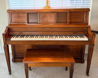 This baldwin piano is finished in a warm wood tone with fluted tapered legs, a full 88-key keyboard, a decorative fretwork panel above the fallboard, and casters at each leg. A matching bench with a lift-top storage seat is included, and a small brass finial ornament sits on top. Tuning and action condition not evaluated. 