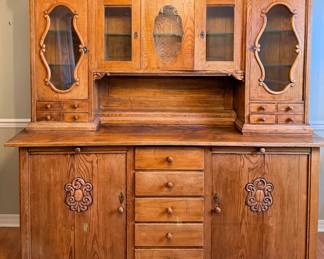 This large hutch has a lower base with four center drawers flanked by two carved-panel cupboard doors with decorative escutcheons, topped by an upper section with two glazed display cabinets, a center carved panel, and four small drawers on either side. An open shelf runs across the middle for display. Substantial storage and display space in one piece, well suited to a dining room or kitchen.