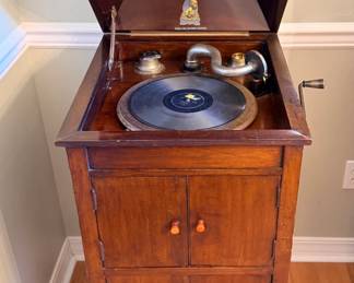 This floor-model wind-up phonograph sits in a wood cabinet with a hinged lid, twin lower doors with turned wood pulls, and a side hand crank. The interior shows the turntable with a record in place, a nickel-finish tone arm and reproducer, and a decorative decal on the underside of the lid. Not tested for play — a striking piece for a collector of early home audio or as a statement furniture piece.