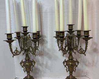 This matched pair of brass candelabra each hold five candles on scrolled arms rising from an ornate urn-form body with a footed base. The casting shows decorative relief work throughout, and the arms carry drip pans at each cup. Candles shown are included as pictured. A dramatic pair for a mantel, dining table, or entryway.