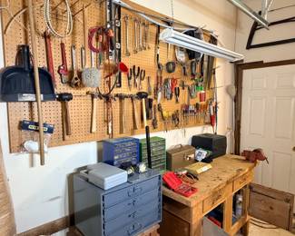 This workshop lot includes a fully loaded pegboard wall of hand tools — wrenches, pliers, hammers, screwdrivers, clamps, saws, extension cords, and long-handled yard tools — along with a wood workbench, a blue metal multi-drawer parts cabinet, a small tool box, a gray storage case, and assorted bench-top items. Quantity is substantial and includes both common and specialty tools. A strong buy for anyone outfitting a shop or garage.