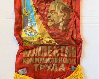 This red banner is printed with a portrait of Lenin alongside an industrial and agricultural scene, with gold Cyrillic lettering reading "КОЛЛЕКТИВ КОММУНИСТИЧЕСКОГО ТРУДА" (Collective of Communist Labor). It finishes with gold bullion fringe along the bottom edge and a small metal medallion at the lower right. A genuine piece of Cold War–era political material for collectors of Soviet artifacts and 20th century history.