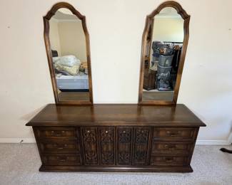 This long dresser has three drawers at each end flanking a center bank fronted by four carved scrollwork panels with round brass knobs, plus antiqued brass drop pulls throughout. Two matching arched mirrors in molded wood frames sit on top. The carving and finish match the armoire and octagonal cabinet listed separately — a strong candidate for buyers assembling the full bedroom grouping.
