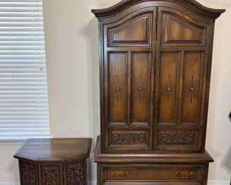 This two-piece furniture lot pairs a tall armoire with an arched bonnet top, four paneled doors with antiqued brass pulls, and two deep lower drawers, along with a matching octagonal side cabinet. Both pieces share the same carved scrollwork panels and dark finish. A substantial bedroom or living room set with generous storage and traditional carved detail.