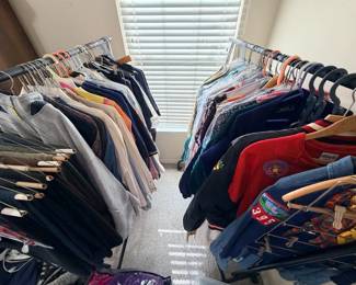 This lot consists of two full rolling garment racks of men's and women's clothing, including sweatshirts, button-down shirts, sweaters, blouses, denim, jackets, and slacks. Several pieces remain in dry-cleaning bags, and the group includes a Quapaw Area Council Scout jacket and other embroidered and patched items. A bulk clothing lot with a wide size and style range — well suited to resellers or anyone shopping volume.