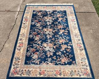 This area rug features a sculpted floral and leaf design in soft rose, cream, and gray against a rich blue field, with a wide cream floral border. The carved pile gives the pattern noticeable depth, and both ends finish with heavy ivory fringe. A classic look for a living room, dining room, or bedroom.