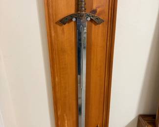 This decorative sword is mounted on a stained wood display plaque and features an ornate cast hilt with a gold-tone pommel, green cabochon accents, and a braided-pattern grip. The blade is marked "Prinz Eisenherz" near the guard, and a small matching miniature sword or letter opener is mounted on the same plaque. A ready-to-hang display piece for fans of decorative swords, medieval-style wall décor, or fantasy collectibles.