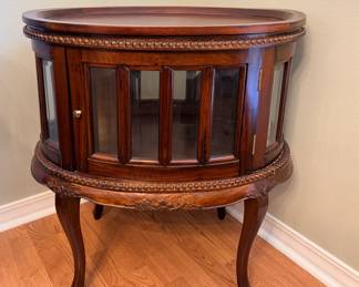 This round accent table features a curved glass display cabinet beneath a wood top, with paneled glass doors that open to interior shelving. Carved rope-twist molding runs around the top and lower edge, and the piece stands on four cabriole legs with carved knee detail. A handsome way to display small collectibles while keeping them dust-free.