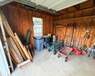 This lot covers the contents of a storage shed: a green folding utility wagon, a red steel wagon, a large group of long-handled yard tools including rakes, shovels, hoes, and edgers hung along the wall, two rolling trash bins, a stack of dimensional lumber and boards, a metal bucket, and assorted small containers on the shelf above. A practical bulk lot for anyone setting up a yard, shop, or property.