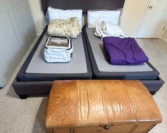 This lot includes an upholstered bed frame in charcoal fabric with a camelback headboard trimmed in nailhead detail, set on two split foundations. A tan leather storage trunk with nailhead trim and a latch closure sits at the foot, usable as a bench, coffee table, or blanket chest. Bedding shown in the photo is included as pictured.