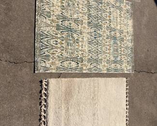 This lot includes a teal and ivory rug in an allover ikat-style geometric pattern and a plain ivory rug with a knotted tassel fringe along one edge. Both are small accent sizes suited to an entry, bedside, or kitchen. Photographed outdoors — colors read true in daylight.
