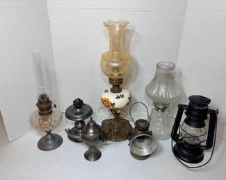This lighting lot includes a large oil lamp with a hand-painted floral font and a fluted amber-toned shade on an ornate metal base, a clear glass oil lamp with a patterned font and chimney, a black metal kerosene barn lantern with a glass globe, a spare ribbed glass shade, and several small pewter-toned metal lamps and finger lamps. Fuel and wick condition not evaluated — sold as found. A nice grouping for display, rustic décor, or emergency lighting.