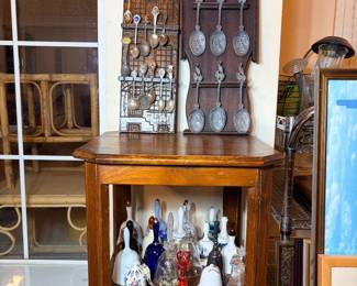 This lot pairs two wall-mounted collector spoon racks — one wood, one wrought iron — filled with souvenir and decorative spoons, with a group of glass and porcelain bells and miniature figural bottles displayed on the shelf below. Colors range across clear, cobalt, amber, and green glass alongside white porcelain pieces. The oak side table shown is included as pictured.