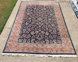 This room-size rug features a dense all-over floral and vine design worked in salmon, ivory, pale blue, and gold across a deep navy field. A wide salmon-toned border with multiple guard stripes frames the field, and both ends finish in ivory fringe. Photographed outdoors in daylight — colors read true, and the pile appears full and even across the surface.