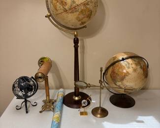 This lot centers on two raised-relief style world globes — a large one on a turned wood floor-height stand with a full brass-tone meridian ring, and a smaller one on a brass-tone disc base. Also included are a wood-and-brass tabletop telescope with a stained-glass shade detail, a black metal open-frame globe on a scrolled iron stand, a brass magnifier on a weighted stand, a small faceted crystal globe on a gold base, and a rolled world map. A well-coordinated grouping for an office, library, or study.