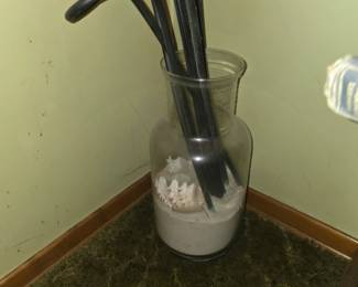 A tall glass jar filled with sand, large sea shells, and black curly rattan diffuser or decorative stems.