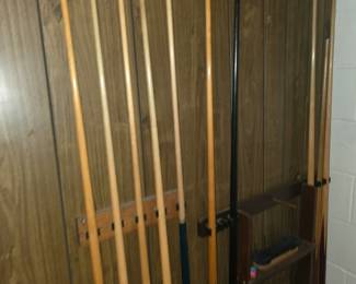 wall-mounted wooden pool cue rack holding several standard two-piece pool cues (sticks), a bridge stick, a wall-mounted floor-standing rack setup with shelves, a brush, and wood paneling on a basement or game room wall.