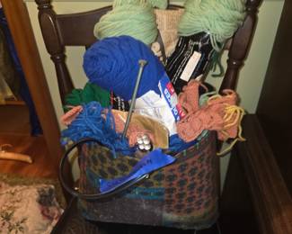 patterned fabric craft or project bag sitting on a chair, filled with an assortment of colorful yarn skeins (including light green, royal blue, and burnt orange/coral), knitting needles, and partially worked projects