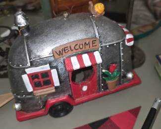 vintage-style decorative mini camper or RV replica (resembling a retro Airstream-style travel trailer) made of resin with a silver metallic finish, red and white striped awning, "WELCOME" sign, and a potted flower accent