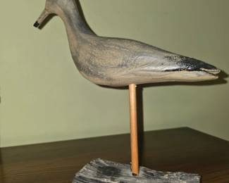 hand-carved and painted wooden shorebird decoy (such as a sandpiper or yellowlegs-style folk art carving) mounted on a vertical wooden rod attached to a rustic wooden or driftwood base