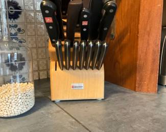 Wüsthof Knife Block Set — Wüsthof branded kitchen knife block set with multiple full-tang stainless steel knives and a wooden storage block. The recognizable Wüsthof shield logo appears on several handles and the block plaque; set includes chef's knife, paring knives, steak knives, honing steel and other utility blades. Good everyday condition with normal handle wear and light blade patina visible in photos; block shows minor surface wear. Compact block measures approx 12in tall and holds a full complement of knives for household cooking.
