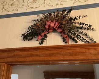 Dried Eucalyptus Doorway Swag — Decorative dried eucalyptus and mixed foliage swag topped with a muted pink ribbon, designed to hang above a doorway or mantel. The arrangement features long eucalyptus stems fanning outward with a dense central cluster of dried filler flowers and a large fabric bow for a cottage or farmhouse look. Estimated overall size fits above a standard doorframe and is lightweight for easy hanging. In good condition with intact foliage and ribbon; naturally fragile as dried botanicals so handle gently.