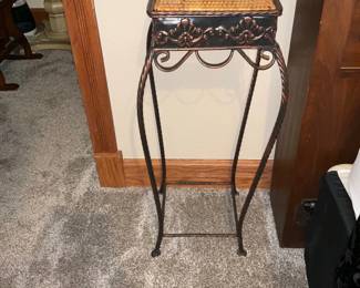 Wrought Iron Plant Stand — Tall wrought-iron plant stand with decorative scrollwork and a woven wicker-top shelf. The metal has a dark finish with copper highlights giving a warm two-tone look; the wicker top shows light wear but remains intact. Sturdy twisted legs and lower stretcher provide stability and a decorative silhouette that works well in entryways or porches. Overall good condition with only surface wear consistent with normal use; remote controls shown are not included.