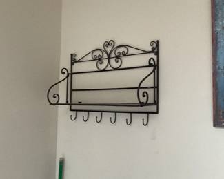 Wrought Iron Wall Shelf with Hooks — Decorative wrought-iron wall shelf featuring scrollwork at the top, two horizontal display shelves and six lower hooks for keys or small items. Solid metal construction with a dark finish shows light surface wear and minor scuffs consistent with normal use but no bent elements visible. Works well as an entryway organizer or kitchen display shelf; overall sturdy and ready to hang with two upper mounting points. Measures compactly and is easy to install on a standard wall.