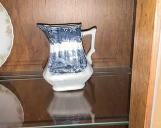 Blue Transferware Cream Pitcher — Small ceramic cream pitcher decorated with blue transferware pastoral scenes and floral border motif. Square-bodied pitcher with a molded handle and spout, glossy white glaze with blue transfer pattern on three sides. Measures approximately 5.5 inches tall, 4.5 inches wide (including spout/handle) and 3.5 inches deep; light and easy to display in a china cabinet. In good condition with light crazing visible in glaze and no obvious chips or cracks.