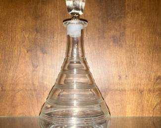 Clear Glass Decanter with Stopper — Clear glass decanter with a faceted stopper and concentric ring decoration makes an elegant addition to a bar or display cabinet. The teardrop-shaped body with stepped horizontal rings and ground-glass stopper suggests mid-century or modern styling and is made entirely of clear glass. Measures approximately 11.5 inches tall with a 5.5 inch widest diameter and is relatively lightweight and easy to handle. In good condition with minor surface wear and a snug-fitting stopper; no visible chips or cracks.