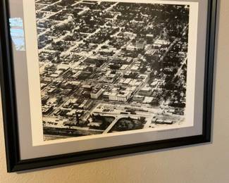 Framed Aerial City Photograph — Framed black-and-white aerial photograph showing a downtown grid with buildings and streets, matted in light gray and housed in a simple black wood frame. The print appears to be a photographic reproduction (possibly labeled in the margin but not fully legible in photos) and is ready to hang; glazing shows light reflection but no obvious cracks. Frame shows minor edge wear consistent with normal hanging; overall good display condition.