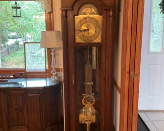 Howard Miller Grandfather Clock — Tall floor-standing grandfather clock featuring a carved bonnet top and brass dial with moon phase complication. The solid wood case houses visible brass weights and lyre-style pendulum behind a full-length glass door, finished in medium brown. Measures approximately 84 inches high, 20 inches wide, and 14 inches deep and is substantial but movable by two people. In good working cosmetic condition with light surface wear to finish consistent with age; clock appears intact though movement function should be verified by buyer.