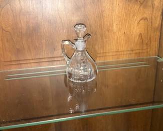 Crystal Glass Oil Cruet with Stopper — Clear pressed crystal cruet with matching faceted stopper, designed for oils or vinegars. The bulbous fluted body, small handle and tapered poured spout give it an elegant, classic look suitable for dining-table use or display in a cabinet. Estimated dimensions fit easily on a shelf; see measurements below for planning transport. In good condition with no visible chips or cracks in the photographed piece; minor surface wear and light shelf dust consistent with use and age.
