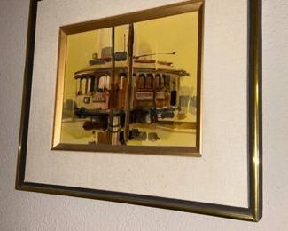 Framed Trolley Watercolor Print — Small framed watercolor-style image depicting a streetcar/trolley with warm yellow background, double-matted in linen-look mat and narrow gold inner frame with brass outer frame. The piece appears to be a reproduction or watercolor study rather than an original signed painting; visible wear is limited to light foxing or age spots on the paper and minor scuffs to the gilt frame. Overall solid wall-ready presentation in a slim profile frame; frame shows small surface dings and one corner separation under the mat as photographed. Useful as vintage-style wall art for a hallway or study.