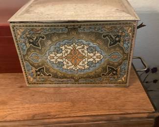 Decorative Metal Storage Tin Box — Decorative hinged metal storage tin featuring ornate floral/medallion printing in blue, gold and cream tones. Rectangular box with hinged lid and side bail-style handles, lined with reflective metal interior suitable for storing household items or games. Approximate overall size and weight make it an easy tabletop or shelf accent for entryways or living rooms. Shows light surface wear, patina and minor dents consistent with age but no holes or major damage.