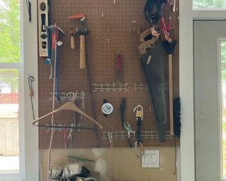 Workshop Pegboard with Hand Tools — Wall-mounted pegboard organizing a small assortment of hand tools and hardware for a home workshop. Visible items include a handsaw, hammers, clamps, jumper cables, tape measure, wire hooks, a small three-drawer parts organizer and assorted small hand tools; board is mounted above a wooden workbench. Overall compact setup is practical for light carpentry and household repairs and is easy to remove for transport. Condition is used with typical wear on tools and surface marks on the pegboard and workbench; tools appear functional but are sold as-is.